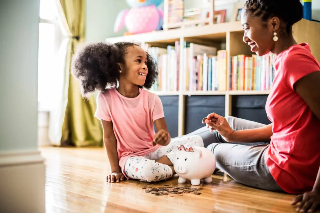 A smiling young girl and an adult sit on the floor, counting coins next to a piggy bank in a bright room with bookshelves and toys in the background.