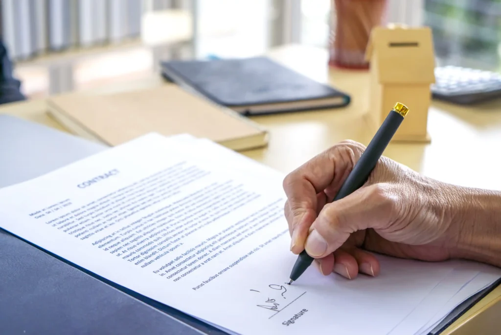 A persons hand holding a pen signs a contract document on a desk, with other paperwork, a notebook, and a small wooden box in the background.
