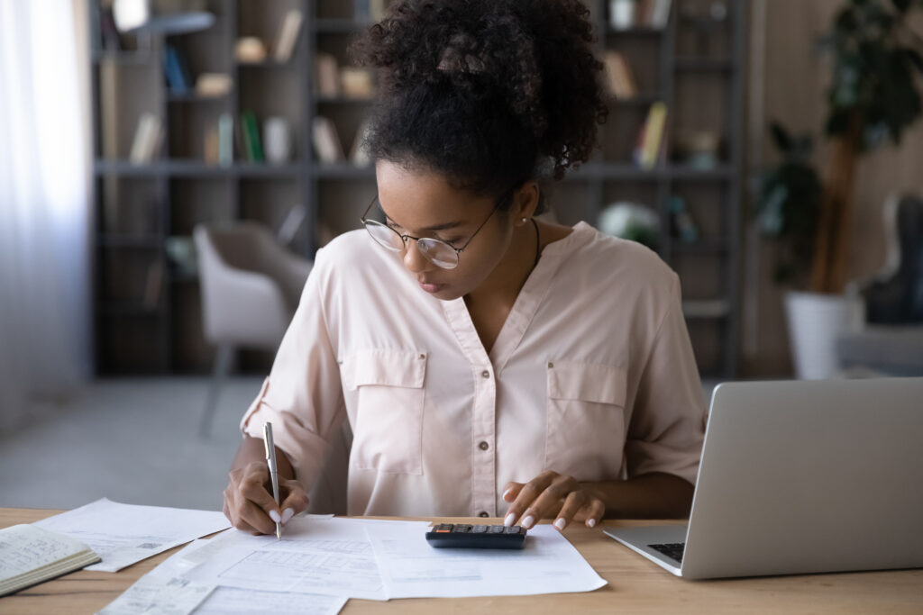 How the 50-30-20 Rule Works Woman seated at a desk reviewing financial documents, writing notes, and using a calculator while working on a laptop in a home office setting.