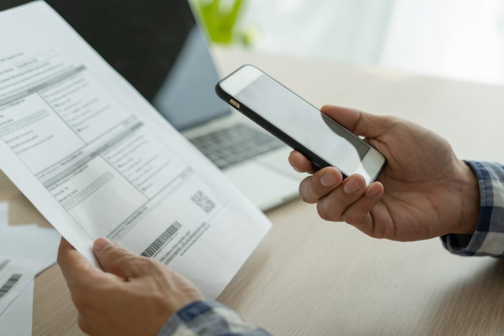 Paying Credit Card With Another Credit Card Person holding a smartphone while reviewing a bill, with a laptop in the background, representing online bill payment or account management.