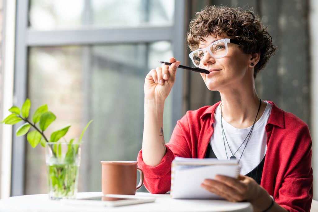 Reasons to Get a Personal Loan Explained Woman sitting by a window, holding a notebook and pen while looking thoughtfully into the distance.
