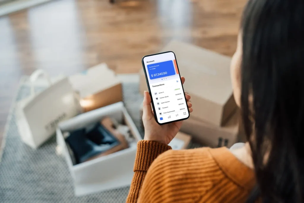 10 Best Online Banks of August 2026 A person in an orange sweater checks their digital wallet balance and transactions on a smartphone, sitting near shopping bags and an open box on a carpeted floor.