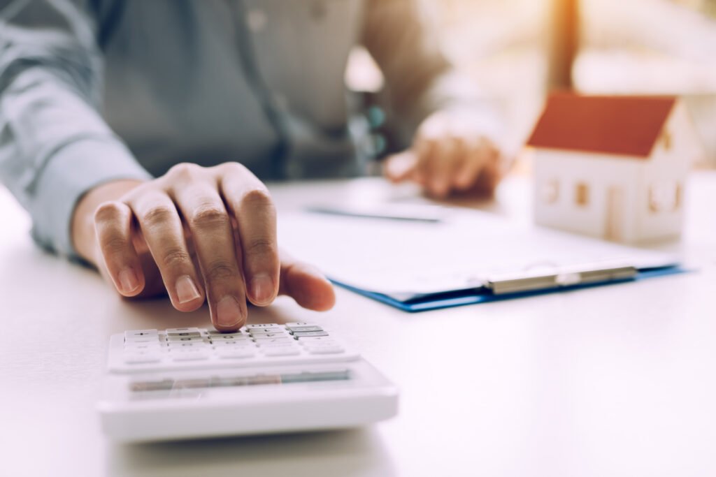 Can I Borrow From My IRA to Buy a House? Person using a calculator beside a miniature house model and mortgage paperwork.