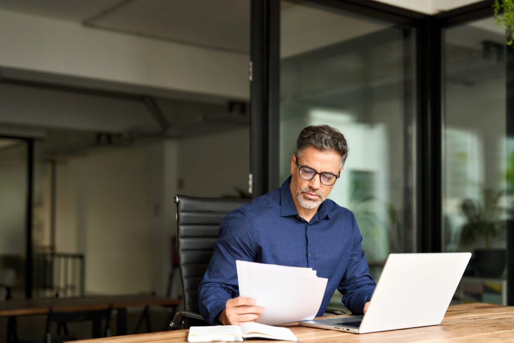 Can You Close a Bank Account With a Negative Balance? Busy mature professional business man using laptop reading document at work.