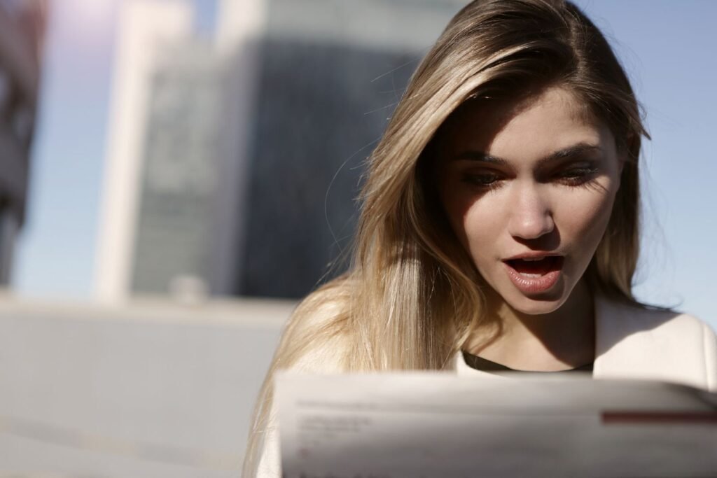 A young woman with blonde hair expresses surprise while reading a newspaper outdoors in daylight.; 2027 Social Security COLA